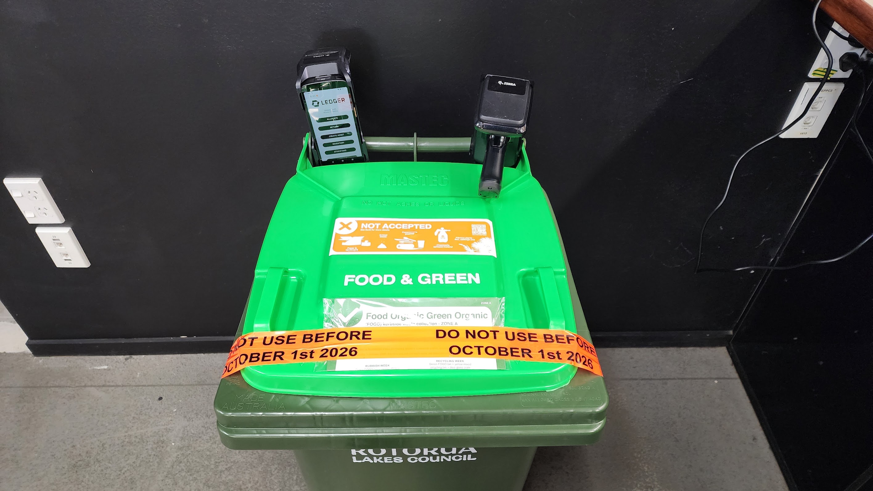 A bin with two handheld scanners placed on top of it. One scanner has a mobile device affixed to it displaying the interface of the Ledger app. The camera angle faces down on the bin, not displaying any other information such as private address.